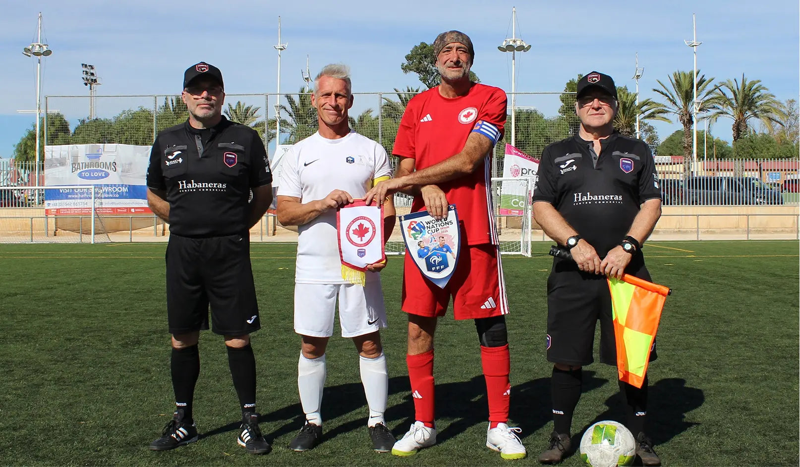 Four men on a soccer field holding penants for France and Canada wearing sports attire. Next to them are referees. In the background are palm trees.