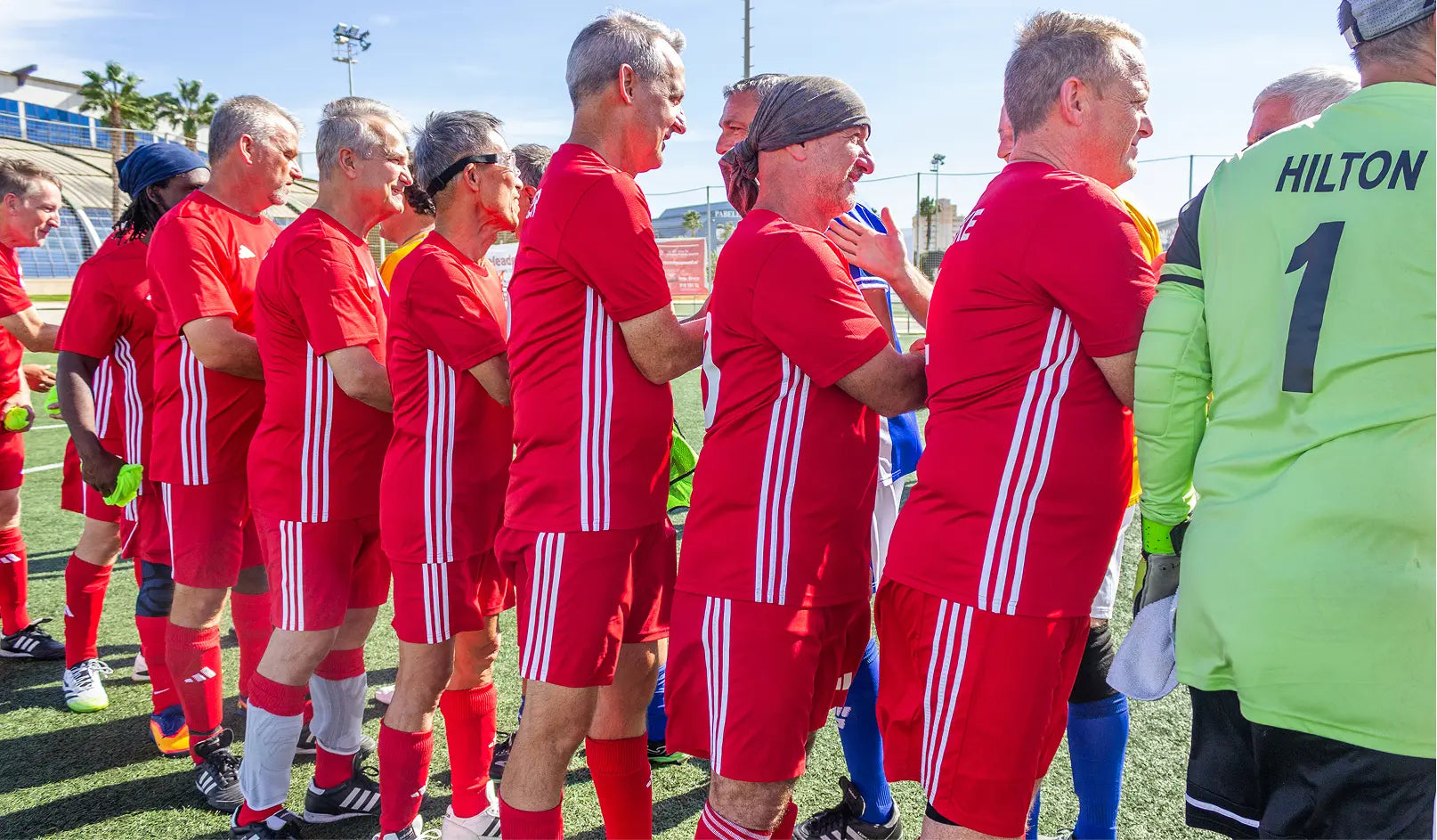 Group of men in red sports uniforms on a soccer field, with one person wearing a green shirt with 'Hilton' branding.