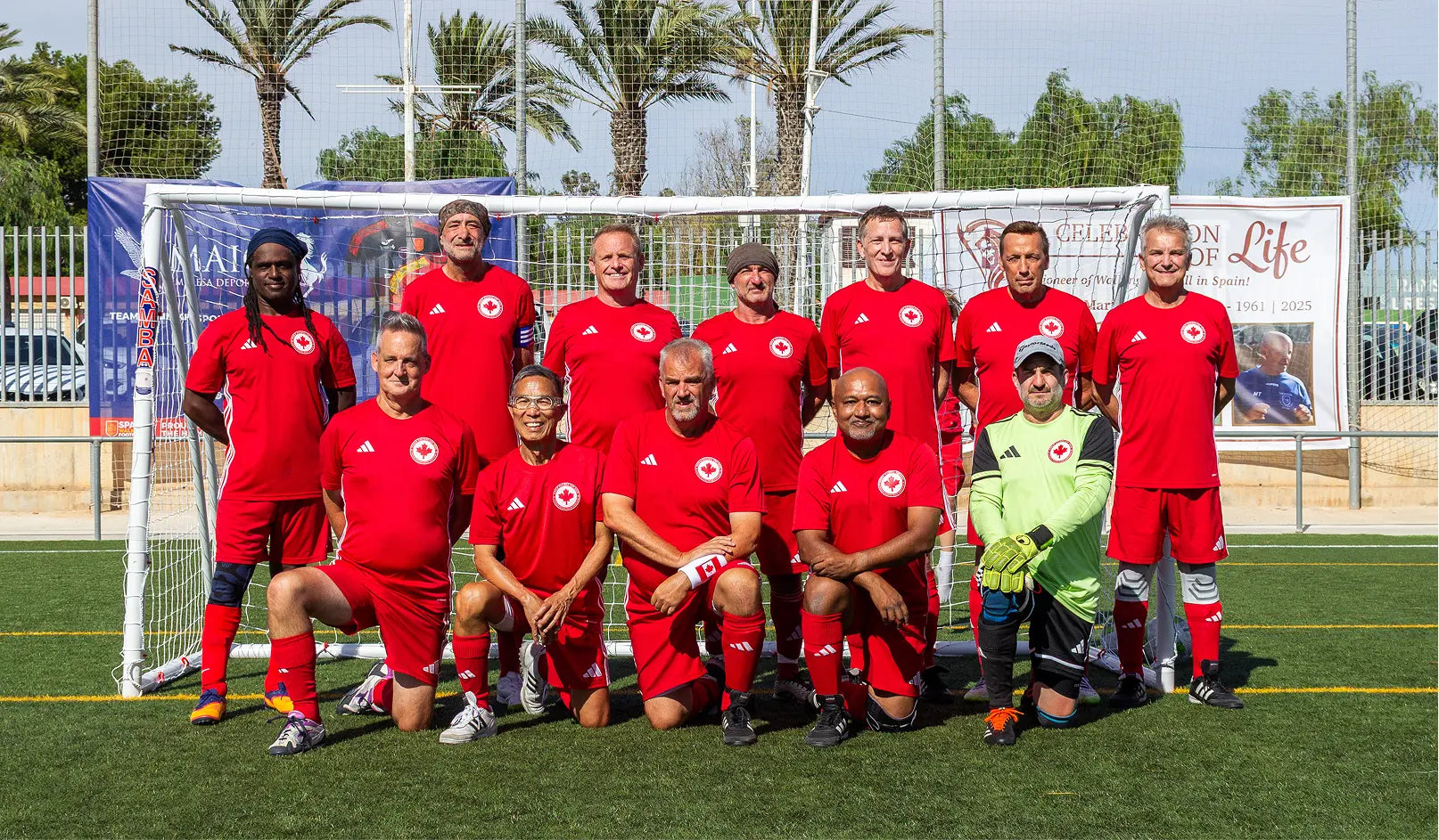 Canada National team soccer players in red uniforms posing on a turf soccer field with palm trees in the background.