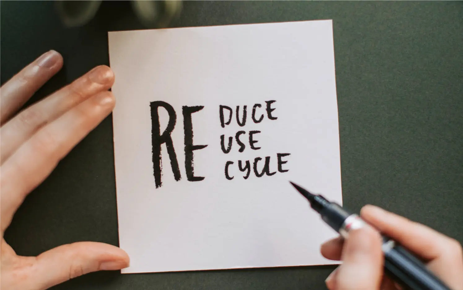 A hand writes a note about sustainability onto a square piece of paper