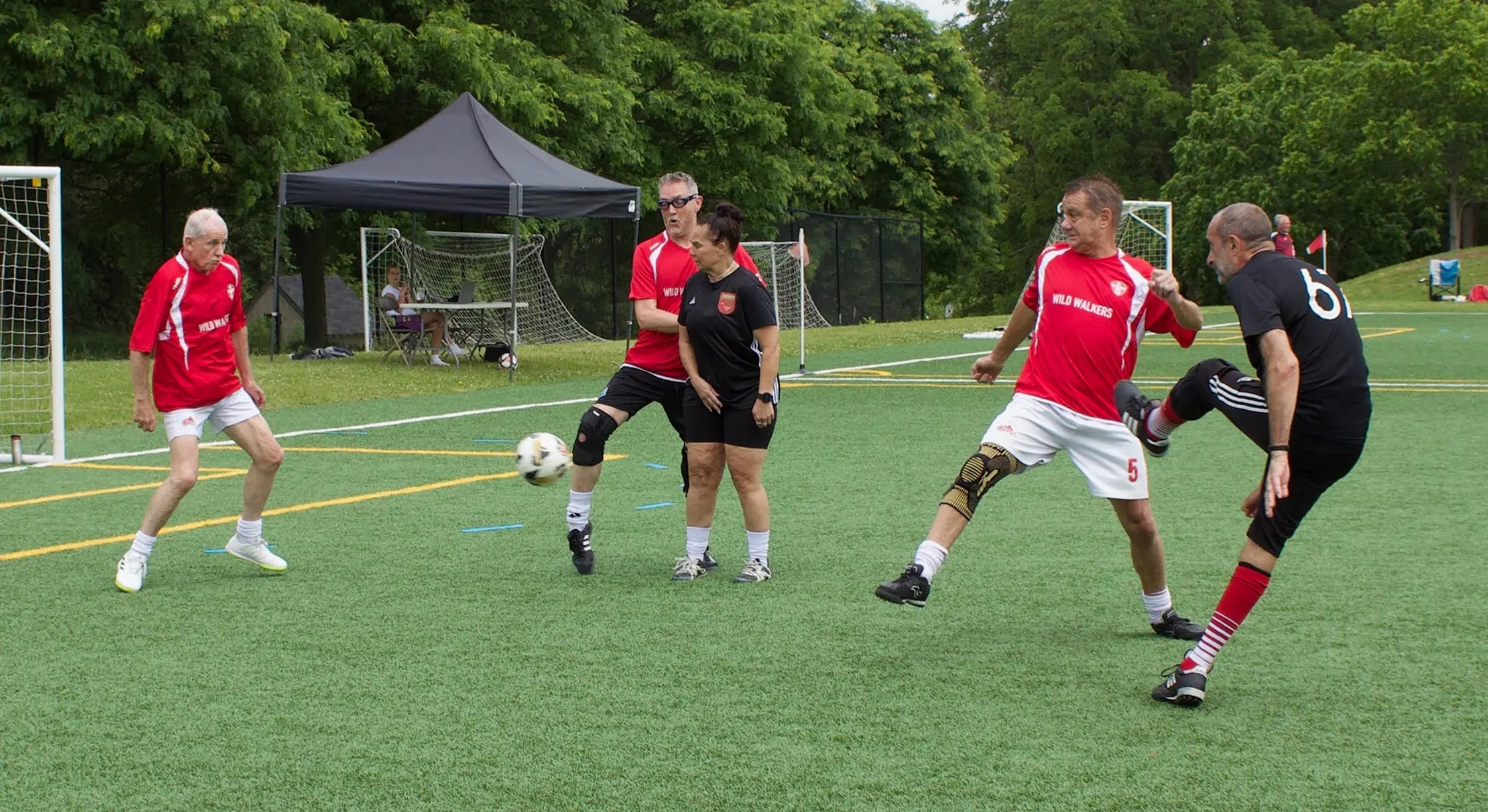 Red team and black team playing soccer on a turf grass field with trees in the background